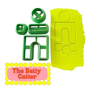 May include: Green plastic cookie cutters in the shape of a person, with a matching impression in a piece of yellow dough. The cutters include a head, torso, and legs. The text "The Betty Cutter" is on a pink background.