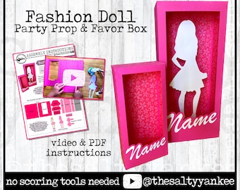 May include: A pink 3D folding template for a Fashion Doll party prop and favor box. The design includes a white silhouette of a doll inside the box, with the word "Name" on the front. Instructions and a video are included.