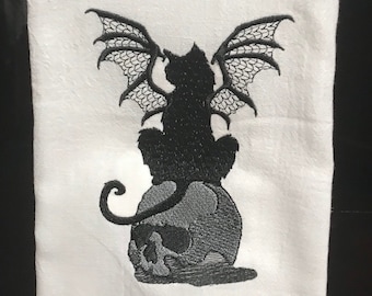 May include: Black and white embroidered kitchen towel featuring a black cat with bat wings sitting on a skull.