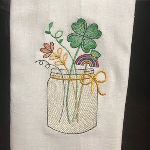 May include: Embroidered kitchen towel with a white background featuring a mason jar filled with green shamrocks, orange flowers, and a rainbow with a leprechaun hat. The jar is tied with a yellow ribbon.