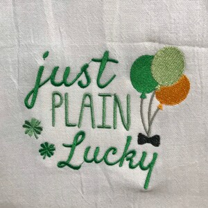 May include: White kitchen towel with green embroidery that reads "just plain lucky" with a green shamrock and a bow tie. There are three green, orange, and yellow balloons with strings.