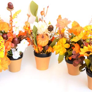 May include: Five small terracotta pots with black rims, each containing a unique artificial floral arrangement featuring autumn-themed elements like orange maple leaves, yellow sunflowers, small pumpkins, acorns, and pinecones.