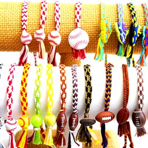 Op de afbeelding: Een display van kleurrijke vriendschapsarmbanden met verschillende sportthema-charms, waaronder honkballen, voetballen, basketbals en tennisballen. De armbanden zijn gemaakt van verschillende gekleurde draden en hebben kwastjes aan de uiteinden.