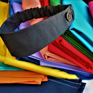 May include: A black fabric headband with a button on the side, sitting on a stack of colorful fabric swatches.