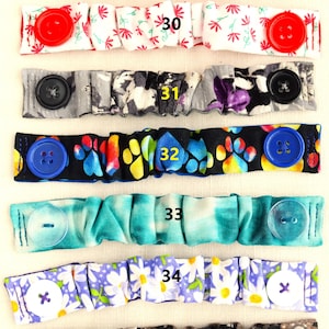 May include: Seven colorful fabric face mask extenders with buttons. The extenders are numbered 29 through 35. The extenders are made of different fabrics with various patterns, including polka dots, flowers, paw prints, tie dye, and daisies.