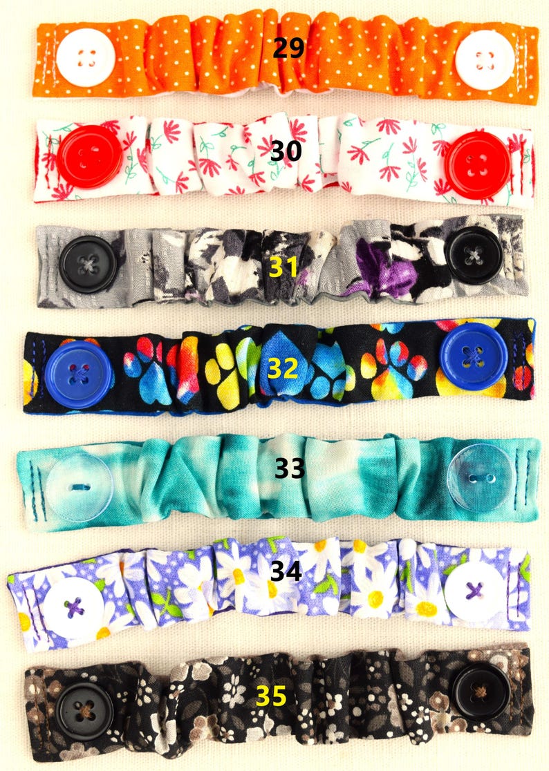 May include: Seven colorful fabric face mask extenders with buttons. The extenders are numbered 29 through 35. The extenders are made of different fabrics with various patterns, including polka dots, flowers, paw prints, tie dye, and daisies.