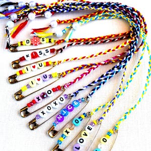 May include: A collection of colorful safety pin charms with braided cords. Each pin features letter beads spelling out words like "KISS," "I LOVE U," and "XOXO." The charms are adorned with beads and tassels.