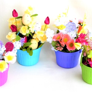 May include: Four colorful Easter basket arrangements. Each basket is a different color: pink, blue, purple, and green. The baskets are filled with an assortment of artificial flowers, decorative eggs, and a bunny cutout.