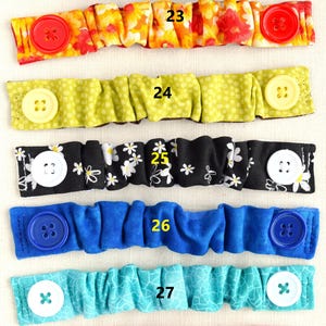 May include: Eight different fabric face mask extenders with buttons. The extenders are in various colors and patterns, including brown with paisley print, orange with floral print, green with polka dots, black with white daisies, blue, turquoise, pink, and white.