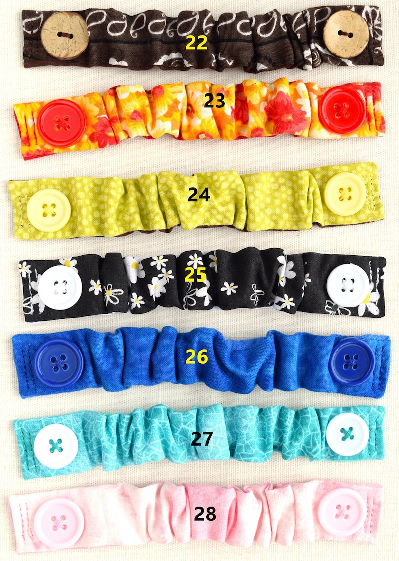 May include: Eight different fabric face mask extenders with buttons. The extenders are in various colors and patterns, including brown with paisley print, orange with floral print, green with polka dots, black with white daisies, blue, turquoise, pink, and white.