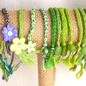 May include: A collection of braided friendship bracelets in shades of green, teal, and yellow, displayed on a burlap-covered stand. Some bracelets feature decorative beads, tassels, and flower charms. The bracelets are handmade and have a bohemian style.