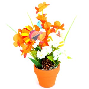 May include: Thanksgiving-themed floral arrangement in a terracotta pot. The arrangement features orange flowers, white blossoms, and a decorative turkey and pumpkin. A pinecone sits at the base of the arrangement. The pot is a vibrant orange color.