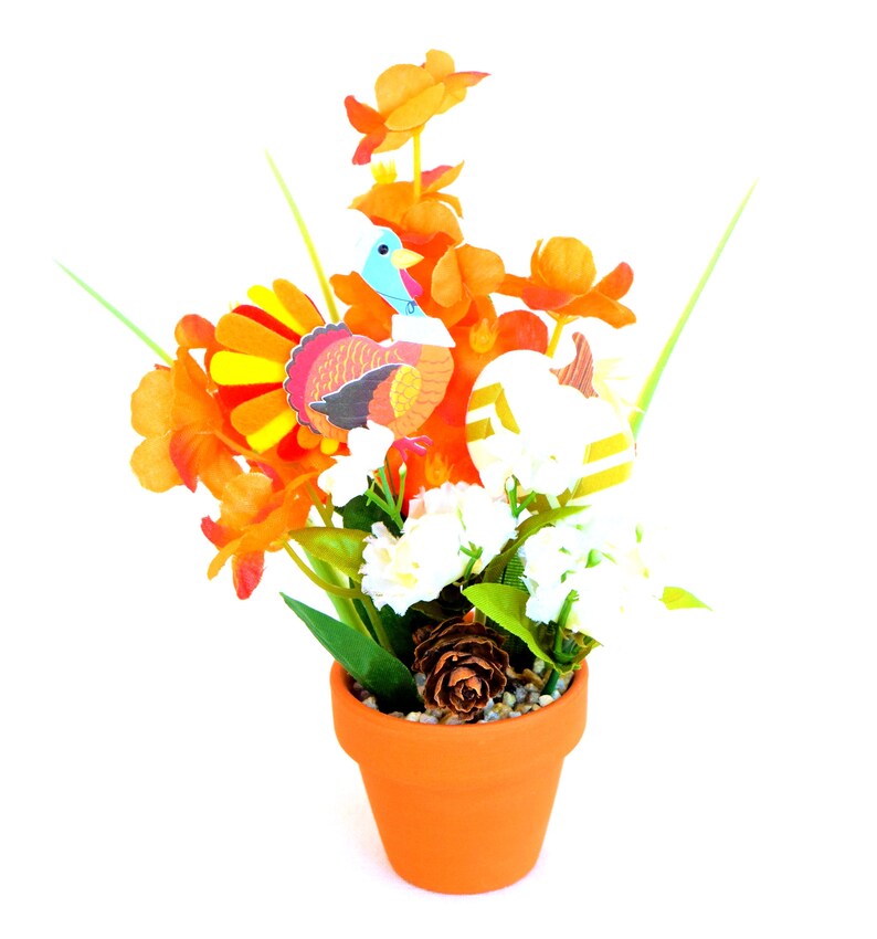 May include: Thanksgiving-themed floral arrangement in a terracotta pot. The arrangement features orange flowers, white blossoms, and a decorative turkey and pumpkin. A pinecone sits at the base of the arrangement. The pot is a vibrant orange color.