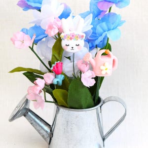 May include: A small metal watering can with a white finish, filled with a bouquet of pink, blue, and white flowers. The flowers are accented with small decorative bunny and watering can charms.