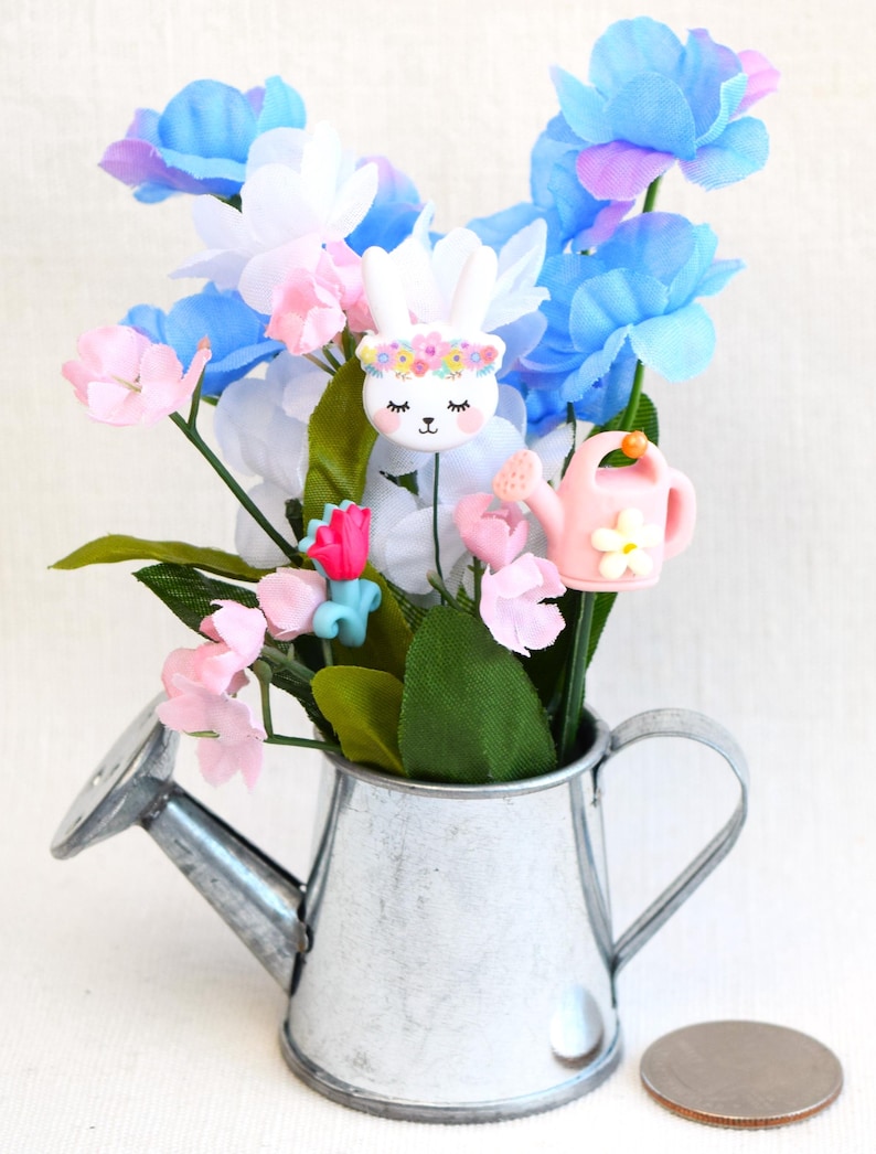 May include: A small metal watering can with a white finish, filled with a bouquet of pink, blue, and white flowers. The flowers are accented with small decorative bunny and watering can charms.