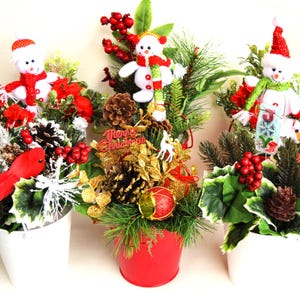 Op de afbeelding: Drie kerstbloemstukken in witte en rode potten. Elk arrangement heeft een sneeuwpop, rode bessen, dennenappels en groen. Eén arrangement heeft een rode vogel en een ander heeft een gouden "Merry Christmas" bord.
