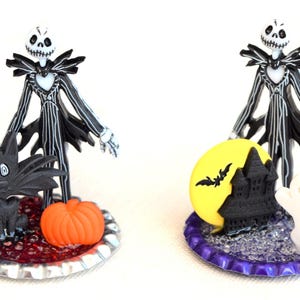 May include: Two Halloween themed bottle cap ornaments featuring a black and white Jack Skellington figure. One ornament features a black cat and a pumpkin, the other features a yellow moon, a black castle, and a white ghost.