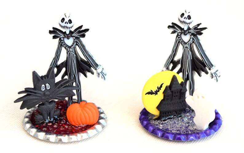 May include: Two Halloween themed bottle cap ornaments featuring a black and white Jack Skellington figure. One ornament features a black cat and a pumpkin, the other features a yellow moon, a black castle, and a white ghost.
