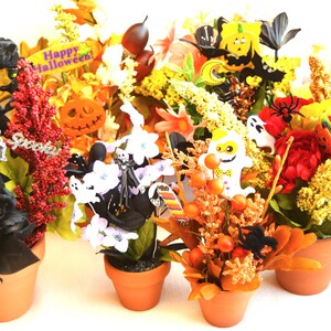 May include: Five Halloween-themed flower arrangements in terracotta pots. Each arrangement features a variety of artificial flowers, leaves, and Halloween decorations, including black roses, orange pumpkins, white ghosts, and black spiders. The sign in the center reads "Happy Halloween!"