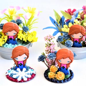 Pu&ograve; includere: Statuine decorative di un personaggio con capelli rossi e abito blu, circondate da fiori artificiali colorati in piccoli secchi di metallo. Una statuina &egrave; su una base decorativa con un motivo a fiocco di neve.