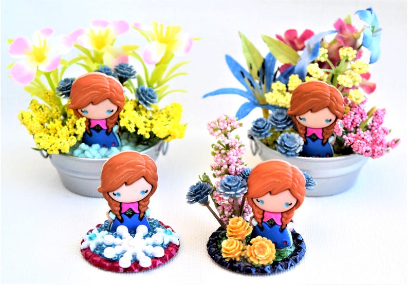 Pu&ograve; includere: Statuine decorative di un personaggio con capelli rossi e abito blu, circondate da fiori artificiali colorati in piccoli secchi di metallo. Una statuina &egrave; su una base decorativa con un motivo a fiocco di neve.