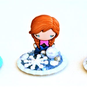 Pu&ograve; includere: Tre figurine in miniatura del film d'animazione Frozen. Olaf il pupazzo di neve, Anna ed Elsa sono raffigurati su basi decorative con dettagli di fiocchi di neve e ghiaccio. Le figurine sono in stile cartone animato con colori vivaci.