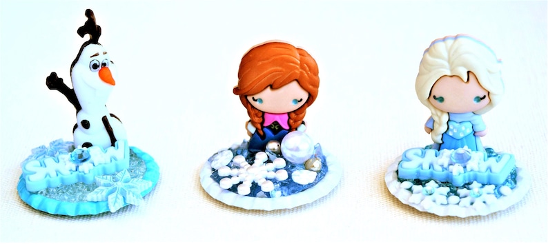 Pu&ograve; includere: Tre figurine in miniatura del film d'animazione Frozen. Olaf il pupazzo di neve, Anna ed Elsa sono raffigurati su basi decorative con dettagli di fiocchi di neve e ghiaccio. Le figurine sono in stile cartone animato con colori vivaci.