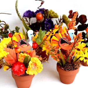 May include: A collection of five small terracotta pots filled with artificial autumn arrangements. Each pot contains a mix of yellow, orange, and red silk flowers, pinecones, berries, and miniature turkey figures with colourful felt tail feathers.