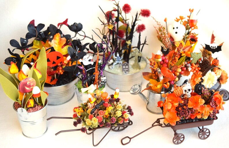 May include: Five Halloween-themed arrangements in various containers, including a wheelbarrow, a watering can, and buckets. The arrangements feature a variety of fall foliage, faux pumpkins, and Halloween decorations, such as ghosts, bats, and skulls.