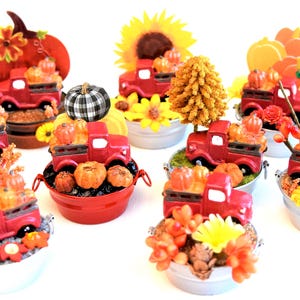 May include: Nine miniature red trucks with white wheels, each carrying miniature pumpkins and surrounded by fall-themed decorations, such as sunflowers, fall leaves, and pine cones. The trucks are arranged in a circle on a white surface.