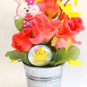 May include: A small galvanized metal bucket filled with a floral arrangement of pink roses, yellow flowers, and green leaves.  The arrangement is decorated with a pink bunny holding an Easter egg, a yellow chick, and a colorful candy.