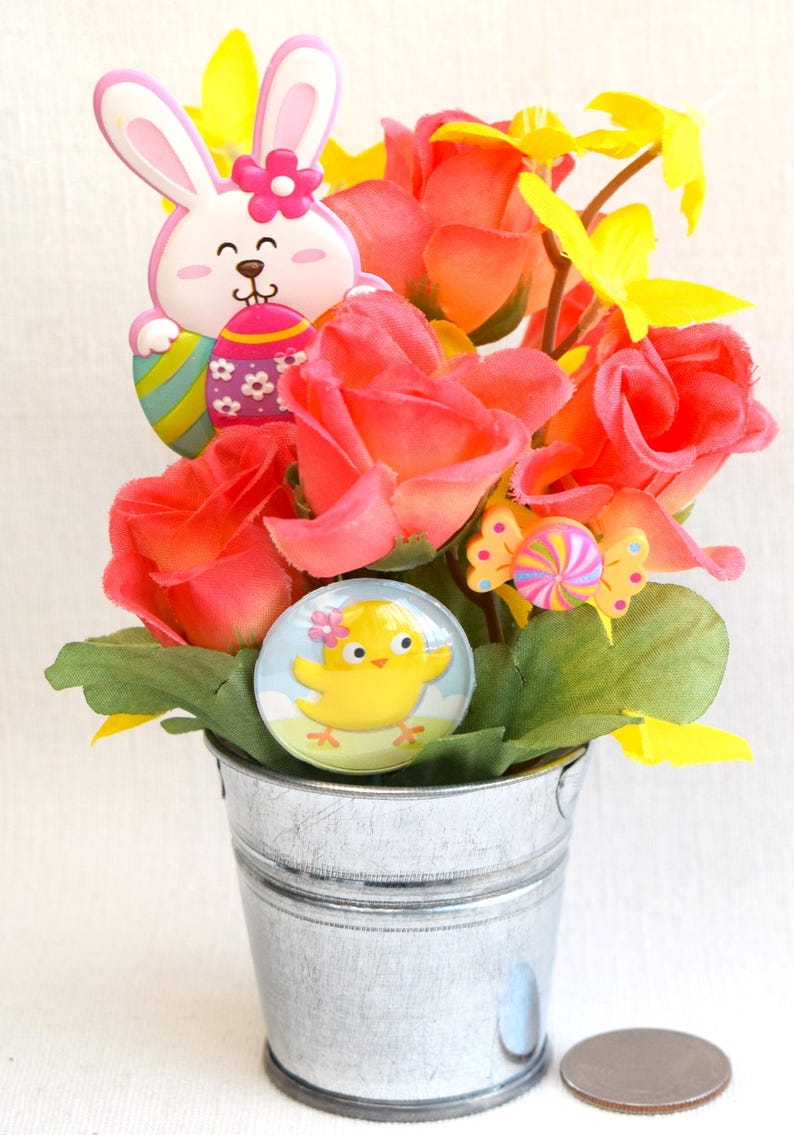 May include: A small galvanized metal bucket filled with a floral arrangement of pink roses, yellow flowers, and green leaves.  The arrangement is decorated with a pink bunny holding an Easter egg, a yellow chick, and a colorful candy.