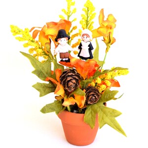May include: A terracotta pot filled with artificial fall foliage, including orange and yellow flowers, green leaves, and pinecones. Two small figurines of a man and woman in traditional pilgrim attire are placed among the flowers.