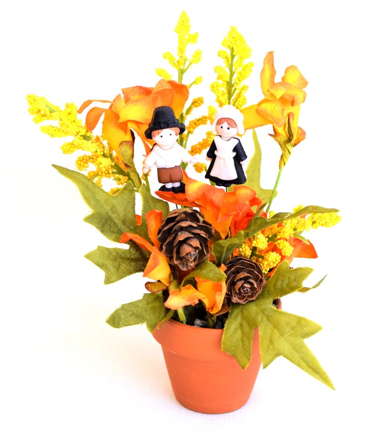 May include: A terracotta pot filled with artificial fall foliage, including orange and yellow flowers, green leaves, and pinecones. Two small figurines of a man and woman in traditional pilgrim attire are placed among the flowers.