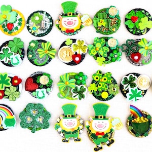 May include: A selection of St Patrick's Day brooches and pins. The pins feature shamrocks, leprechauns, pots of gold, and rainbows. The brooches are in shades of green, gold, and red, with glitter and other decorative elements.