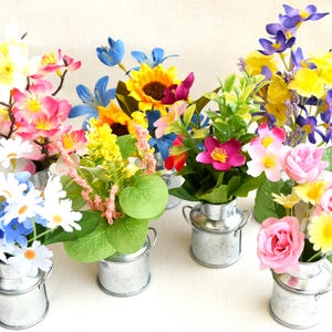 Może przedstawiać: Sześć małych galwanizowanych metalowych dzbanków na mleko z uchwytami, każdy wypełniony innym układem kolorowych sztucznych kwiatów. Kwiaty obejmują różowy, żółty, niebieski, biały i fioletowy.