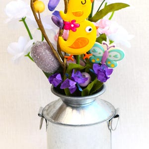 May include: A small galvanized metal milk can vase filled with a floral arrangement. The arrangement includes purple, white, and pink flowers, along with two yellow birds and a blue butterfly.