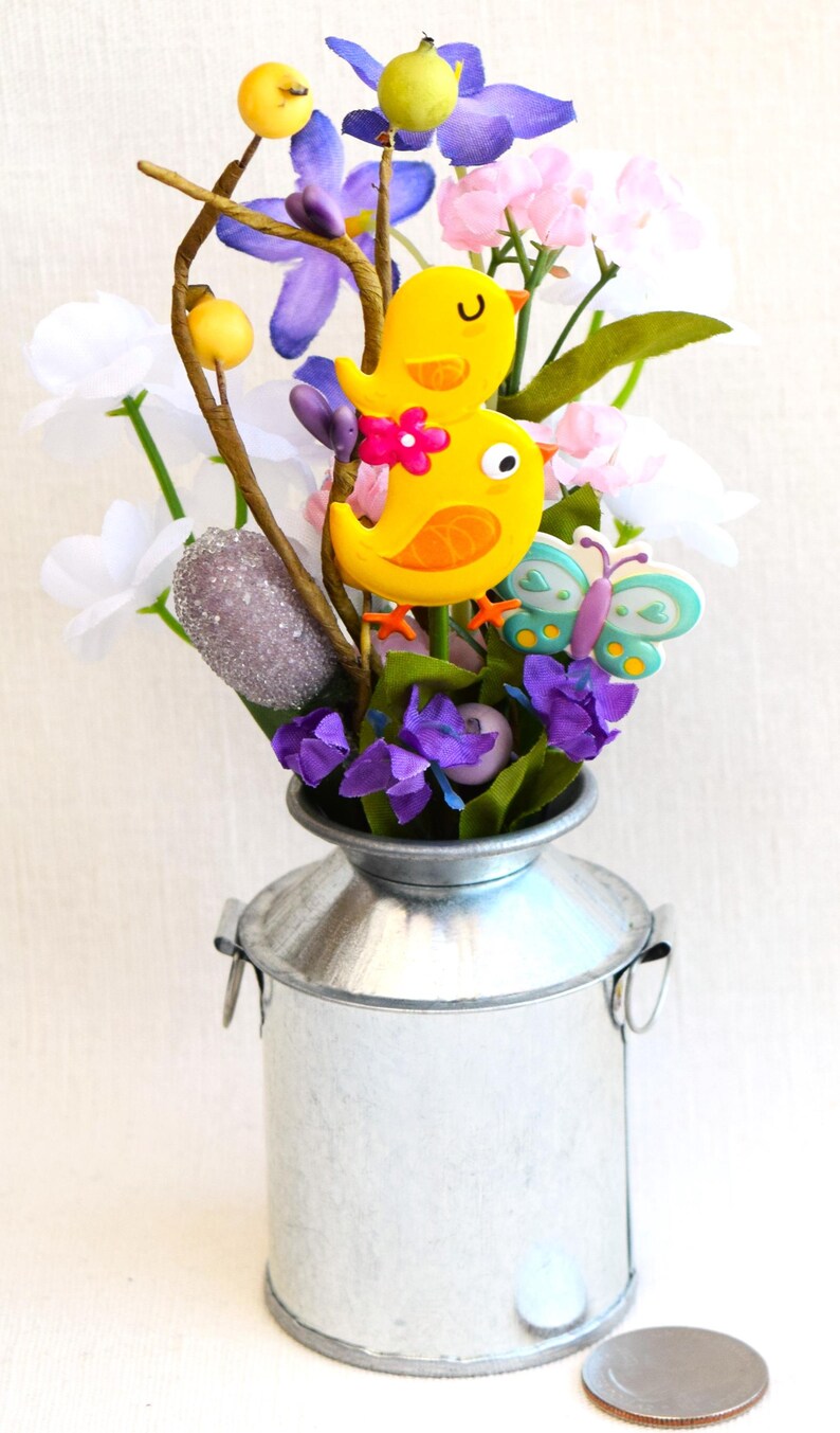 May include: A small galvanized metal milk can vase filled with a floral arrangement. The arrangement includes purple, white, and pink flowers, along with two yellow birds and a blue butterfly.