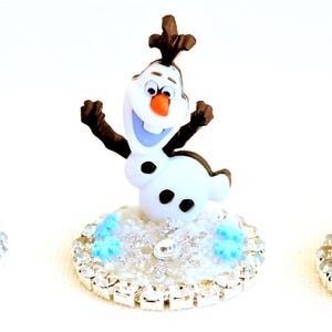 Pu&ograve; includere: Tre figurine dei personaggi del film d'animazione Frozen. Anna con i capelli rossi, Olaf il pupazzo di neve ed Elsa con i capelli bianchi, ciascuna su una base color argento con un motivo a fiocco di neve. Le figurine sono in stile cartone animato.