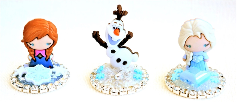 Pu&ograve; includere: Tre figurine dei personaggi del film d'animazione Frozen. Anna con i capelli rossi, Olaf il pupazzo di neve ed Elsa con i capelli bianchi, ciascuna su una base color argento con un motivo a fiocco di neve. Le figurine sono in stile cartone animato.