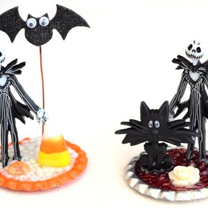 May include: Two Halloween themed figurines, one of Jack Skellington with a bat and candy corn, the other of Jack Skellington with a black cat. Both figurines are on decorative bases.