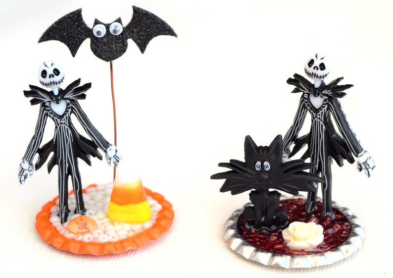 May include: Two Halloween themed figurines, one of Jack Skellington with a bat and candy corn, the other of Jack Skellington with a black cat. Both figurines are on decorative bases.