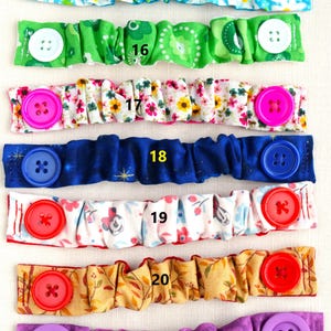 May include: Seven colorful fabric face mask extenders with buttons. The extenders are numbered 15 through 21. The extenders are made of different fabrics with various patterns, including floral, polka dot, and geometric designs. The buttons are in various colors, including white, pink, blue, red, and purple.