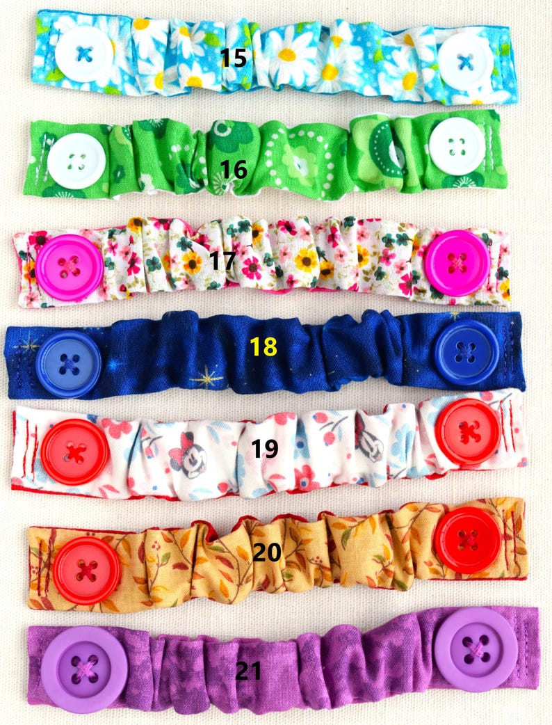 May include: Seven colorful fabric face mask extenders with buttons. The extenders are numbered 15 through 21. The extenders are made of different fabrics with various patterns, including floral, polka dot, and geometric designs. The buttons are in various colors, including white, pink, blue, red, and purple.