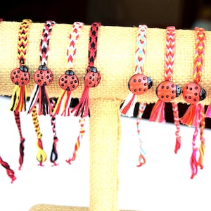 May include: A display of braided friendship bracelets with ladybug charms. The bracelets feature various color combinations, including red, black, yellow, and white. Each bracelet has a tassel and is displayed on a beige stand.