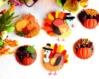 May include: A collection of colorful fall-themed decorative pins. The pins feature pumpkins, turkeys, and fall foliage in shades of orange, yellow, and brown. Some turkeys wear pilgrim hats. These pins are perfect for fall and Thanksgiving.