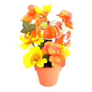 May include: A terracotta pot filled with vibrant orange and yellow flowers. A decorative pumpkin and leaf are attached to the arrangement. The flowers are in full bloom, creating a cheerful autumn display.