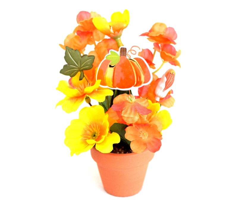 May include: A terracotta pot filled with vibrant orange and yellow flowers. A decorative pumpkin and leaf are attached to the arrangement. The flowers are in full bloom, creating a cheerful autumn display.