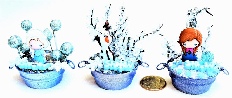 Pu&ograve; includere: Tre composizioni decorative in miniatura a tema Frozen in piccoli secchielli di metallo blu chiaro. Ognuna presenta una statuina di personaggio, neve finta e accenti glitterati. Una moneta &egrave; inclusa per riferimento alle dimensioni.