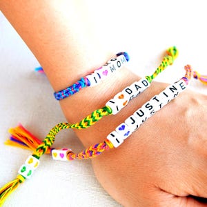 Op de afbeelding: Drie kleurrijke vriendschapsarmbanden met gepersonaliseerde berichten. Eén armband zegt "I MOM" met een hartje, een andere zegt "I DAD" met een hartje en de derde zegt "JUSTINE". De armbanden zitten om een pols.
